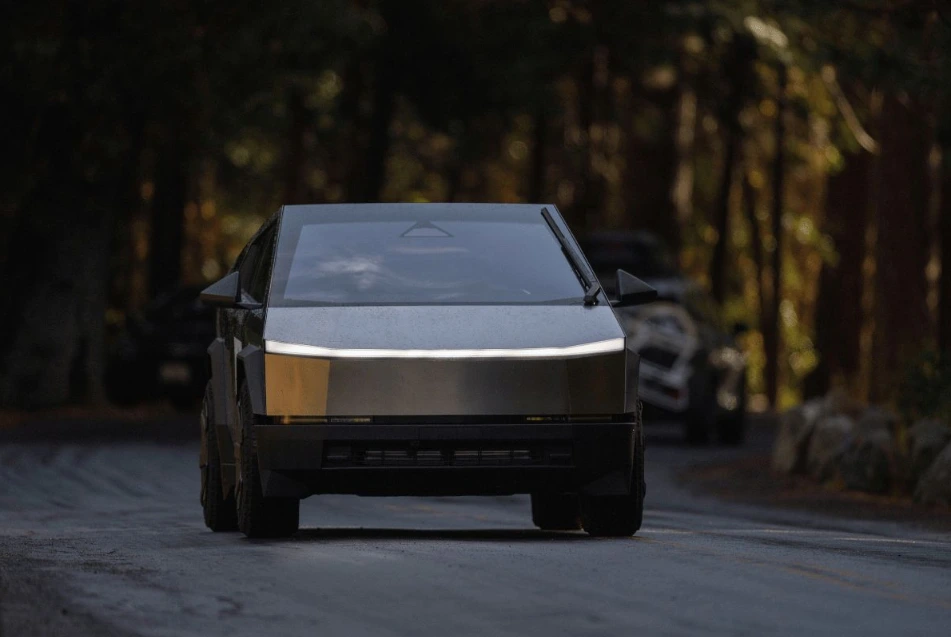 A dramatic, low-light, head-on view of the gray Tesla Cybertruck driving down a paved, winding road surrounded by a dense, dark forest. The truck's angular, futuristic design and horizontal headlight strip are prominent.