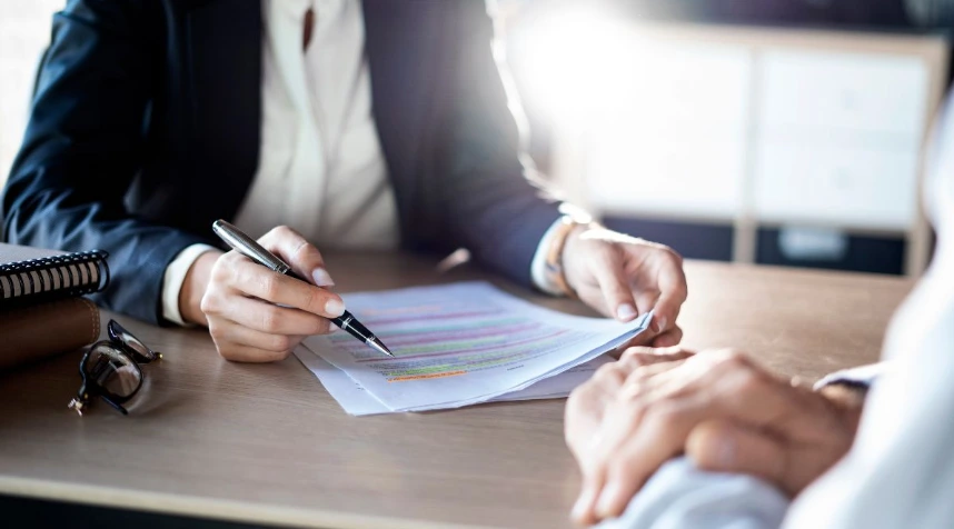 A close-up shot of a female attorney in a dark suit pointing with a pen to a specific highlighted section of a legal document or contract on a wooden desk. A pair of glasses and a notebook are visible in the foreground, with bright light coming from the ba
