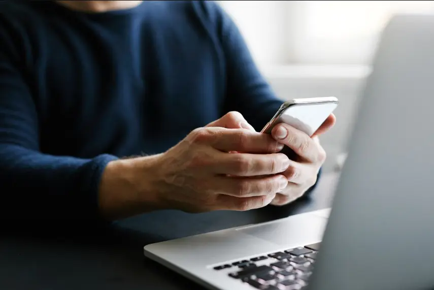 A close-up shot of a person's hands holding and using a silver smartphone, while seated at a dark desk in front of an open laptop. The person is wearing a dark blue long-sleeve shirt.