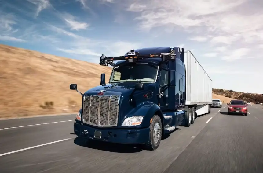A dark blue Peterbilt semi-truck, equipped with a visible autonomous driving sensor rig on its roof, drives on a highway next to a bare, dusty hillside. The truck is hauling a white trailer.