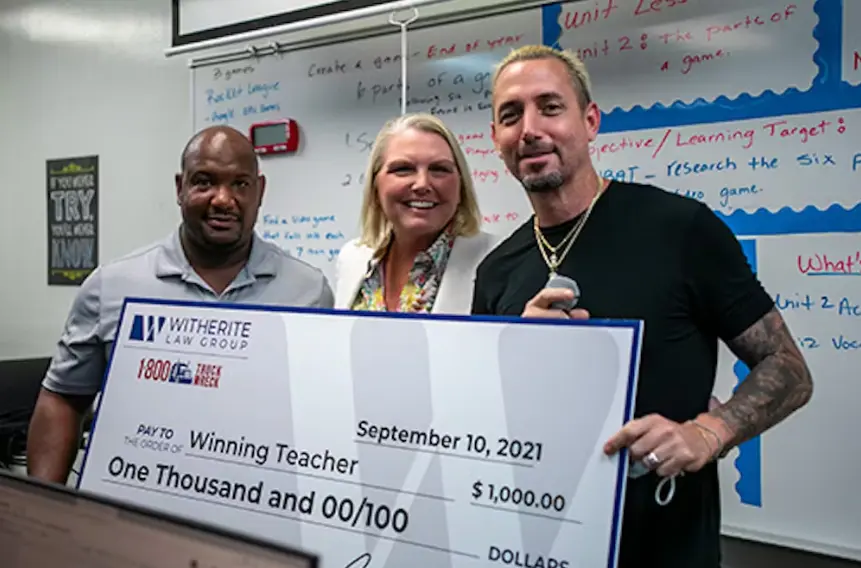Amy Witherite, a blonde woman, smiles while standing between a Black man in a gray polo and a man with tattoos and a beard, who is holding a giant check. The check, dated September 10, 2021, is for "$1,000.00" to the "Winning Teacher" from Witherite Law Gr