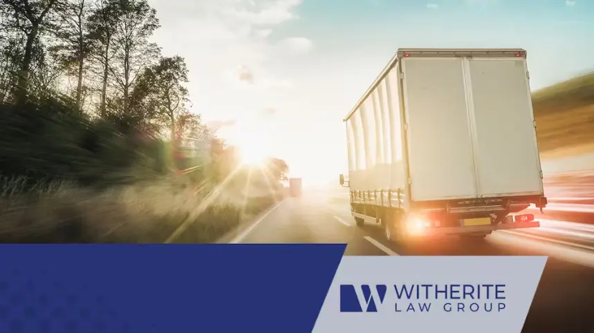 A rear-view, action shot of a white box truck driving down a tree-lined highway at high speed, with bright sunlight and lens flare from the setting or rising sun. The image uses motion blur to convey speed, and the Witherite Law Group logo is overlaid in t