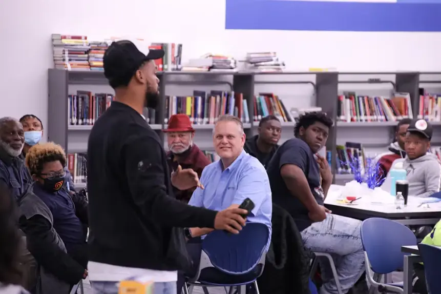 A young man in a black jacket and baseball cap stands with his back to the camera, presenting to a diverse audience seated in a library or classroom setting. Bookcases filled with books are visible behind the seated group, and a smiling older white man in 