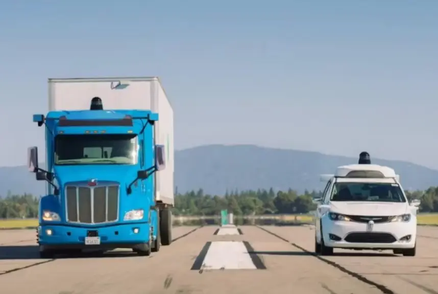 A side-by-side shot of a bright blue semi-truck and a white minivan, both equipped with autonomous driving sensor rigs on their roofs, driving on a paved, deserted road or proving ground with mountains in the background.