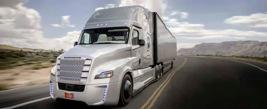 A futuristic, silver Freightliner Cascadia semi-truck with a matching trailer drives on a winding road through a mountainous, desert landscape under a cloudy sky. The truck features modern LED daytime running lights.