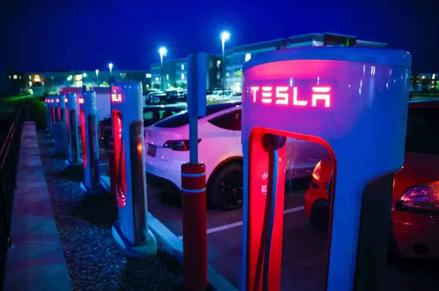 A line of Tesla Supercharger stations illuminated with glowing red lights at night. A white Tesla vehicle is visible charging in the background, parked beneath the bright overhead streetlights of an apartment or commercial complex.