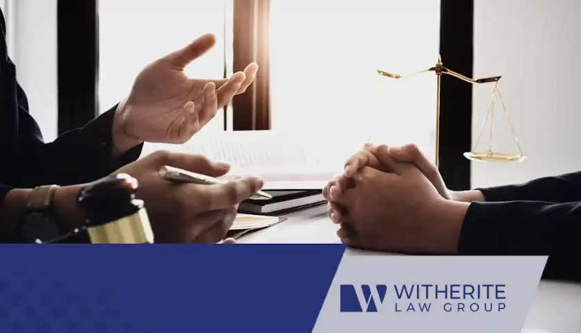 A lawyer is seated at a table, gesturing with his hand and holding a pen during a client consultation. The client's hands are clasped on the table. A small set of golden scales of justice and a gavel are visible on the left, and the Witherite Law Group log