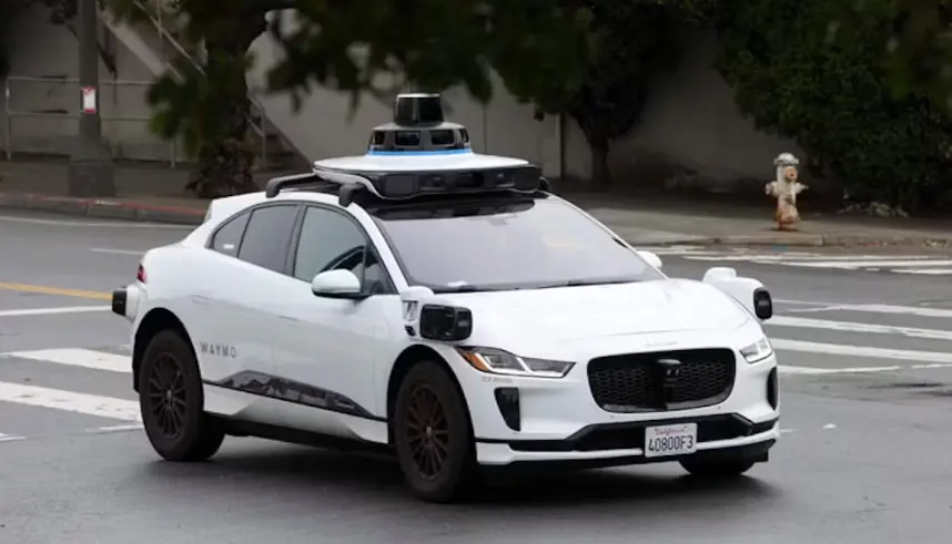 A white Waymo self-driving Jaguar I-PACE SUV, clearly identifiable by the sensor dome on its roof and "WAYMO" decal on the side, is driving on a city street. A California license plate is visible on the front bumper.