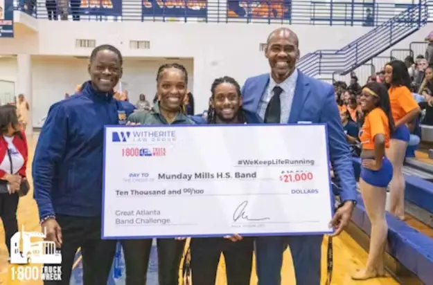Four smiling people—two adults and two younger individuals—hold a large novelty check for "$21,000" made out to the "Munday Mills H.S. Band" for the Great Atlanta Band Challenge. The check features the Witherite Law Group and 1-800-TRUCK-WRECK logos, and c