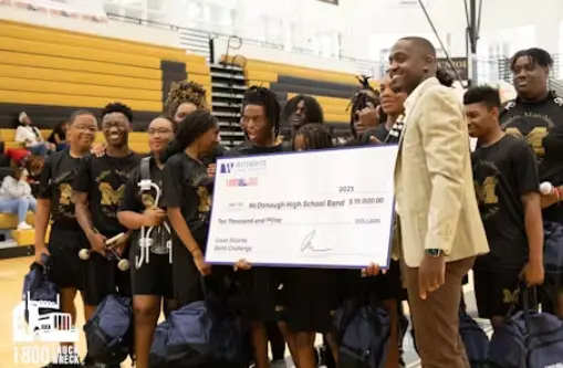 A group of McDonough High School band members and a man in a tan suit (on the right) pose in a gymnasium, holding a large novelty check for "$10,000.00" for the Great Atlanta Band Challenge. The check features the Witherite Law Group and 1-800-TRUCK-WRECK 