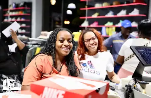 Two smiling women are standing behind a Nike shoe box at a retail store counter. The woman on the right is wearing an Adidas t-shirt and glasses. Shelves of sneakers and other customers are visible in the background.