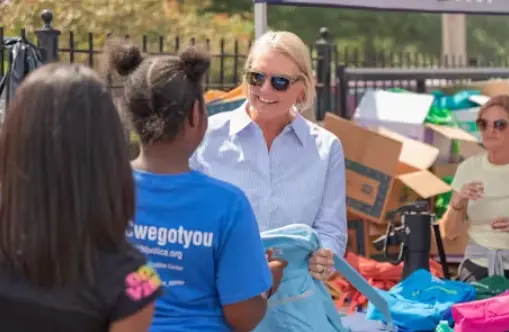 Amy Witherite, a blonde woman wearing a striped shirt and sunglasses, smiles while handing a light blue backpack to a young girl in a blue shirt at an outdoor community event. Many boxes and bags are stacked in the background.