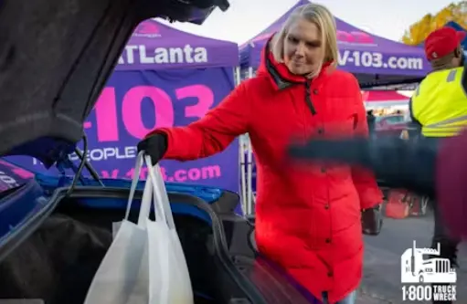 Amy witherite wearing a red puffy winter jacket and black gloves places white plastic bags into the open trunk of a blue car. She is at an outdoor event with a purple tent in the background featuring text for "103" and "Atlanta." The 1-800-TRUCK-WRECK logo