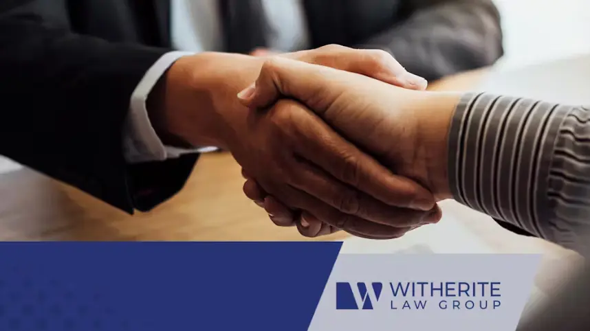 A close-up shot of two people shaking hands across a table, symbolizing a business agreement or partnership. One person is wearing a dark suit, and the other is wearing a striped shirt. The Witherite Law Group logo is overlaid in the lower right corner.