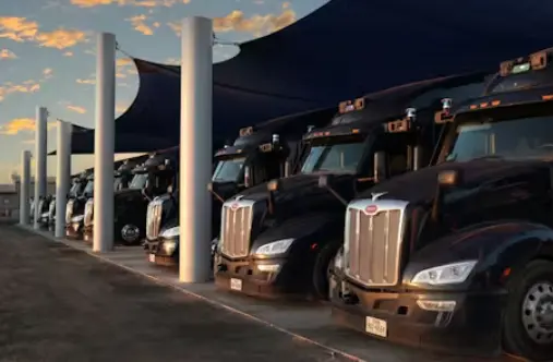 A line of black Peterbilt semi-trucks with matching black cabs are parked closely together under a dark fabric awning supported by concrete columns. The photo is taken at ground level with a focus on the front of the trucks, against a bright, warm sunset s
