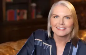 A headshot of Amy Witherite, smiling, wearing a dark navy blue silk blouse with a light geometric pattern. She is seated in a dimly lit, traditional setting, possibly on a leather sofa, with a bookshelf in the blurred background.