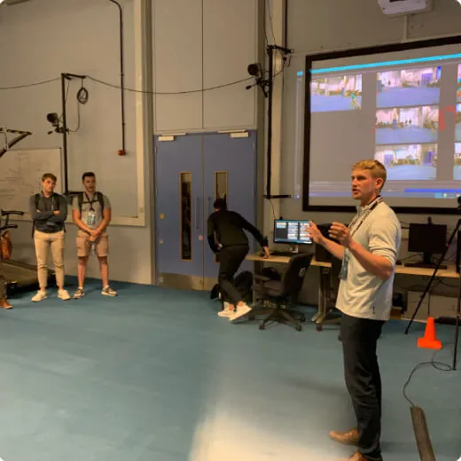 A group of people in a classroom setting, with one man presenting in front of a screen while others observe.