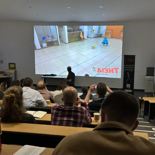 A presenter stands in front of an audience, showcasing a large screen with a demonstration in a spacious auditorium.