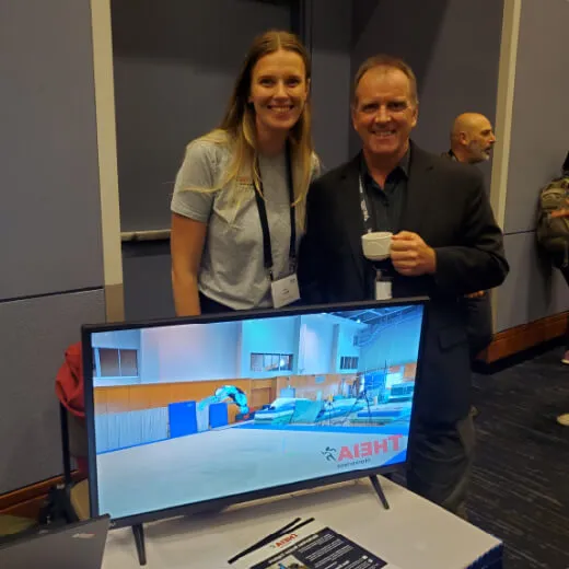 A man and woman stand next to a TV displaying a gymnasium scene. The man holds a cup, smiling, while the woman stands beside him.