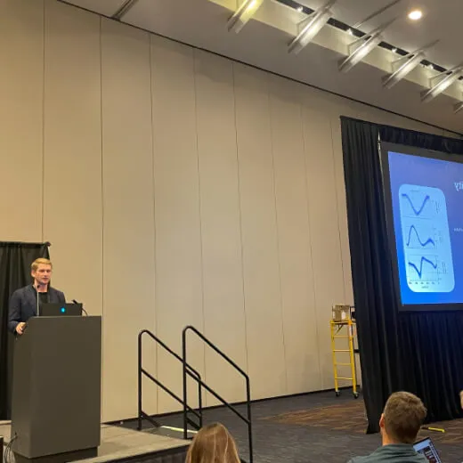 A speaker presents on stage, next to a large screen displaying graphs, with an audience visible in the foreground.