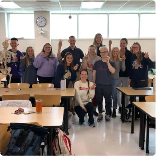 Group of students and a teacher posing with peace signs in a classroom, with desks and a skeleton model visible.