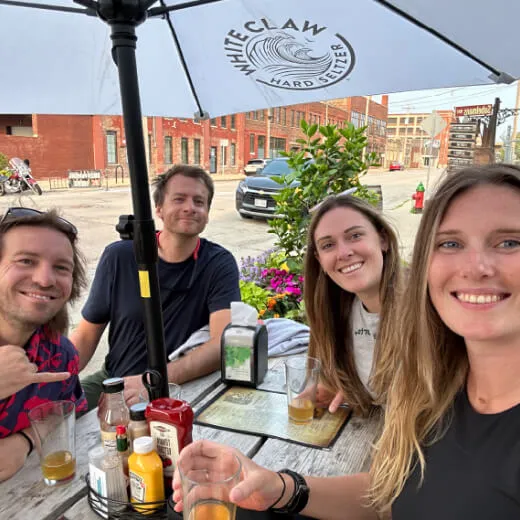 Four friends enjoying drinks at an outdoor table under a White Claw umbrella, with a scenic urban background.
