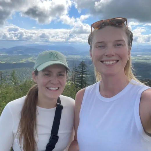 Two women smiling for a selfie outdoors, with a scenic landscape of mountains and trees in the background under a cloudy sky.