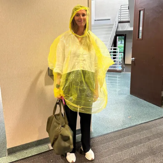 Person in a yellow poncho stands indoors, holding a bag, near a doorway and stairs. Bright, summery attire contrasts with indoor setting.