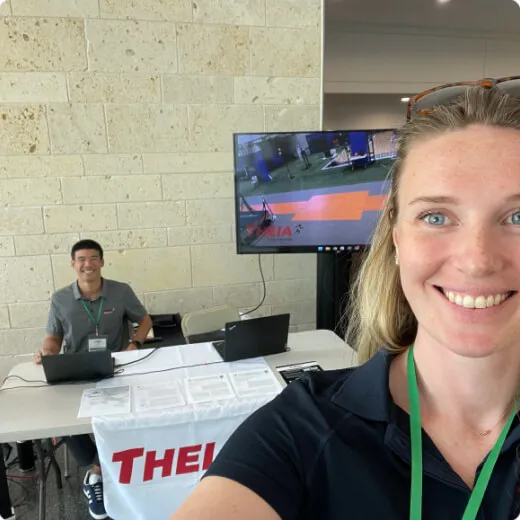 A woman smiles for a selfie at a booth with a man behind a table and a monitor showing an exercise demonstration.