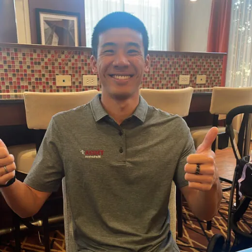 A smiling person with short hair in a gray polo shirt gives a thumbs-up. They are seated indoors with a colorful background.