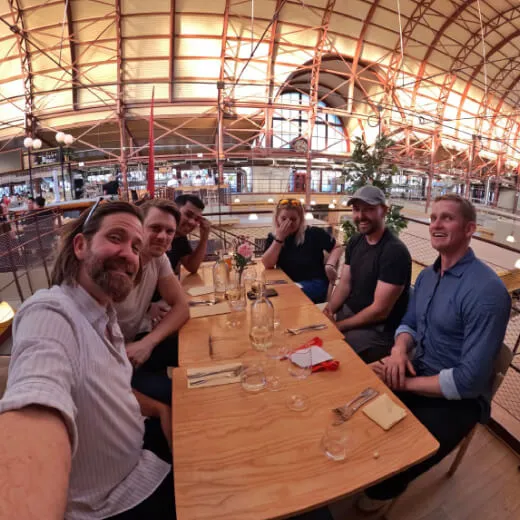 A group of six people enjoys a meal at a lively restaurant with a high ceiling and plants on the table.