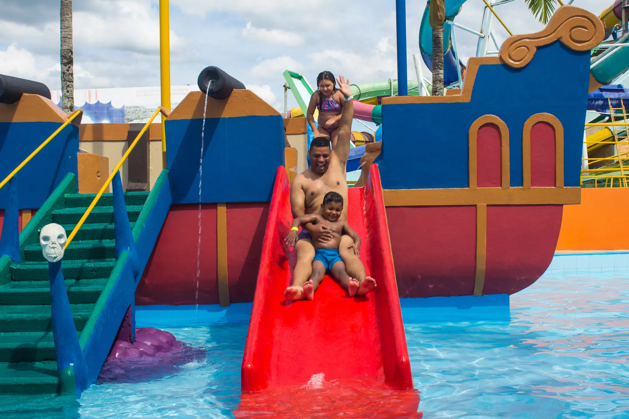 Crianças sentadas na borda de uma piscina, perto de uma grande rampa aquática que sai de uma casa na árvore com tema de abelhas, com um prédio colorido e árvores ao fundo.
