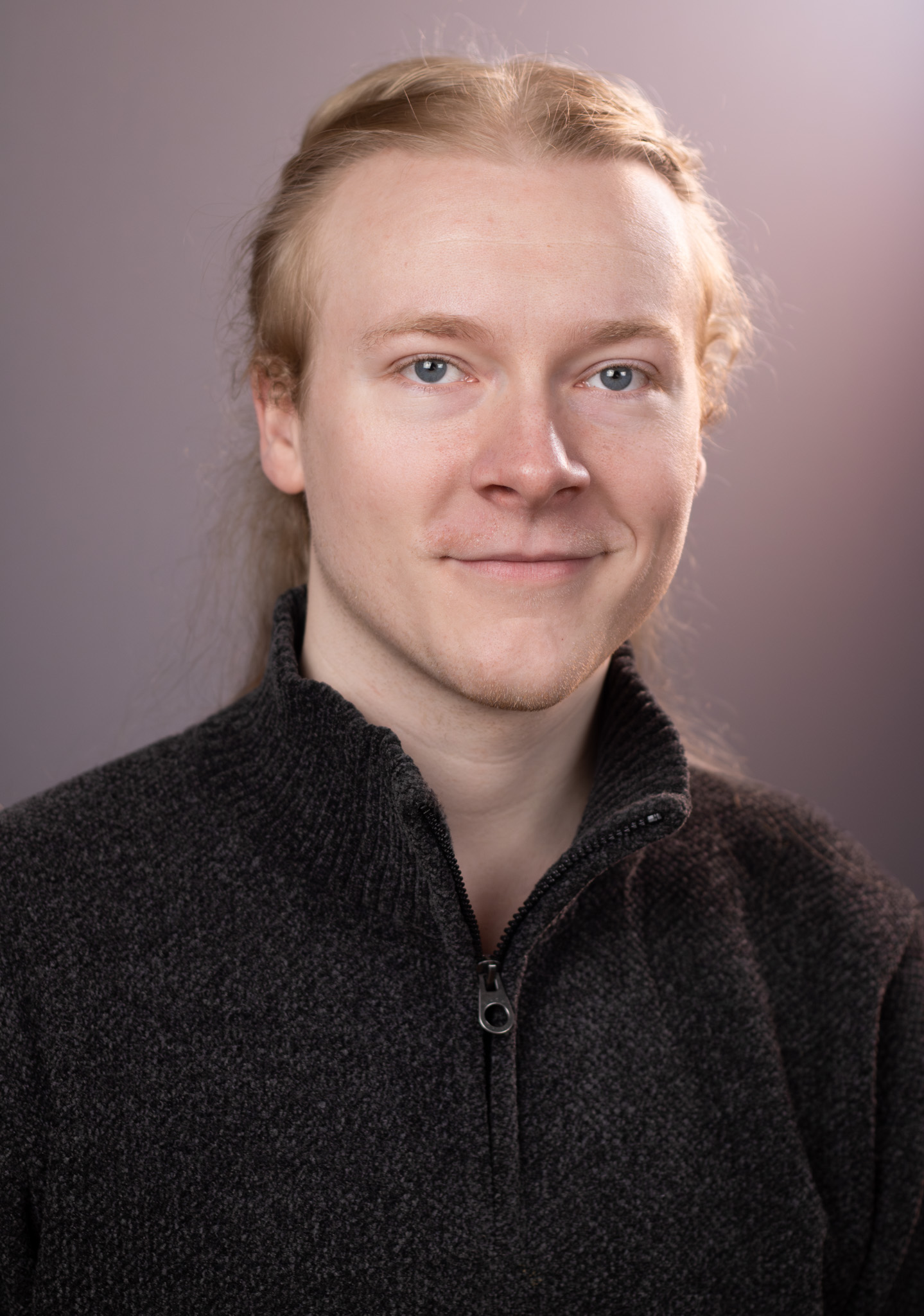 John's headshot. He gazes at the camera with a relaxed smile, and his blond hair is split into two braids, which disappear behind his head.