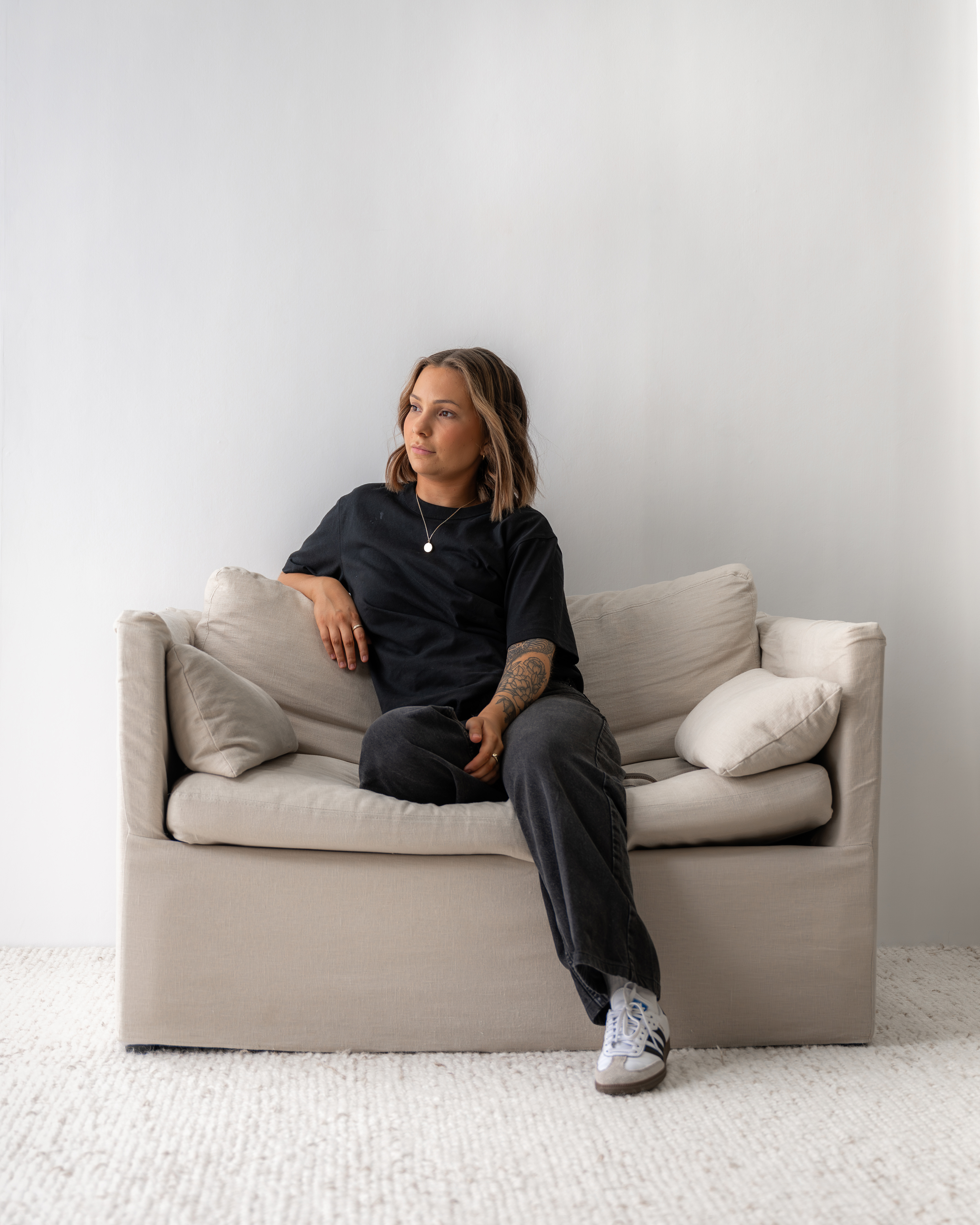 Woman with tattoo on her arm sitting on a beige couch looking to the side in a minimalistic room.
