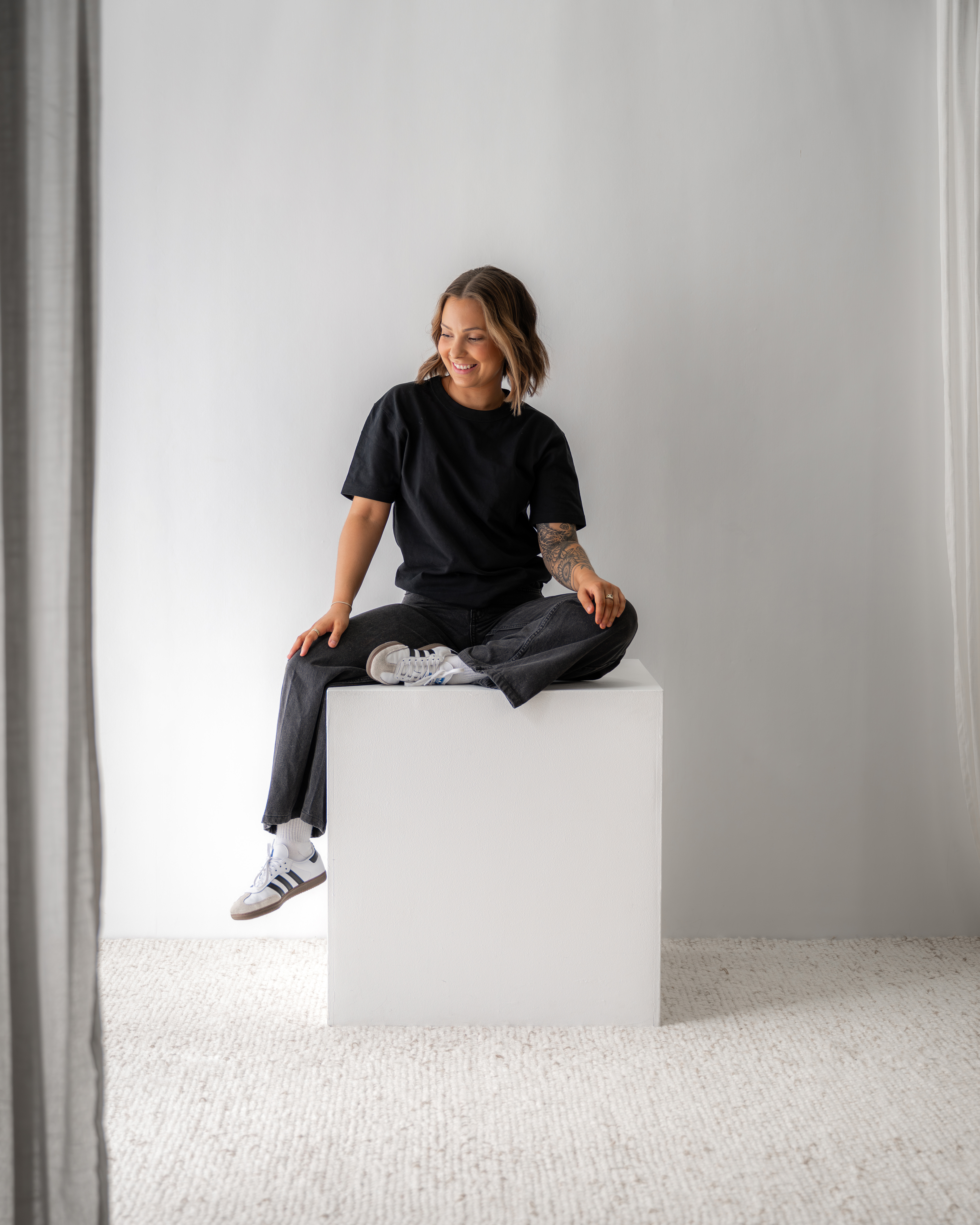 Smiling woman with tattooed arm sitting cross-legged on a white cube in a bright room.
