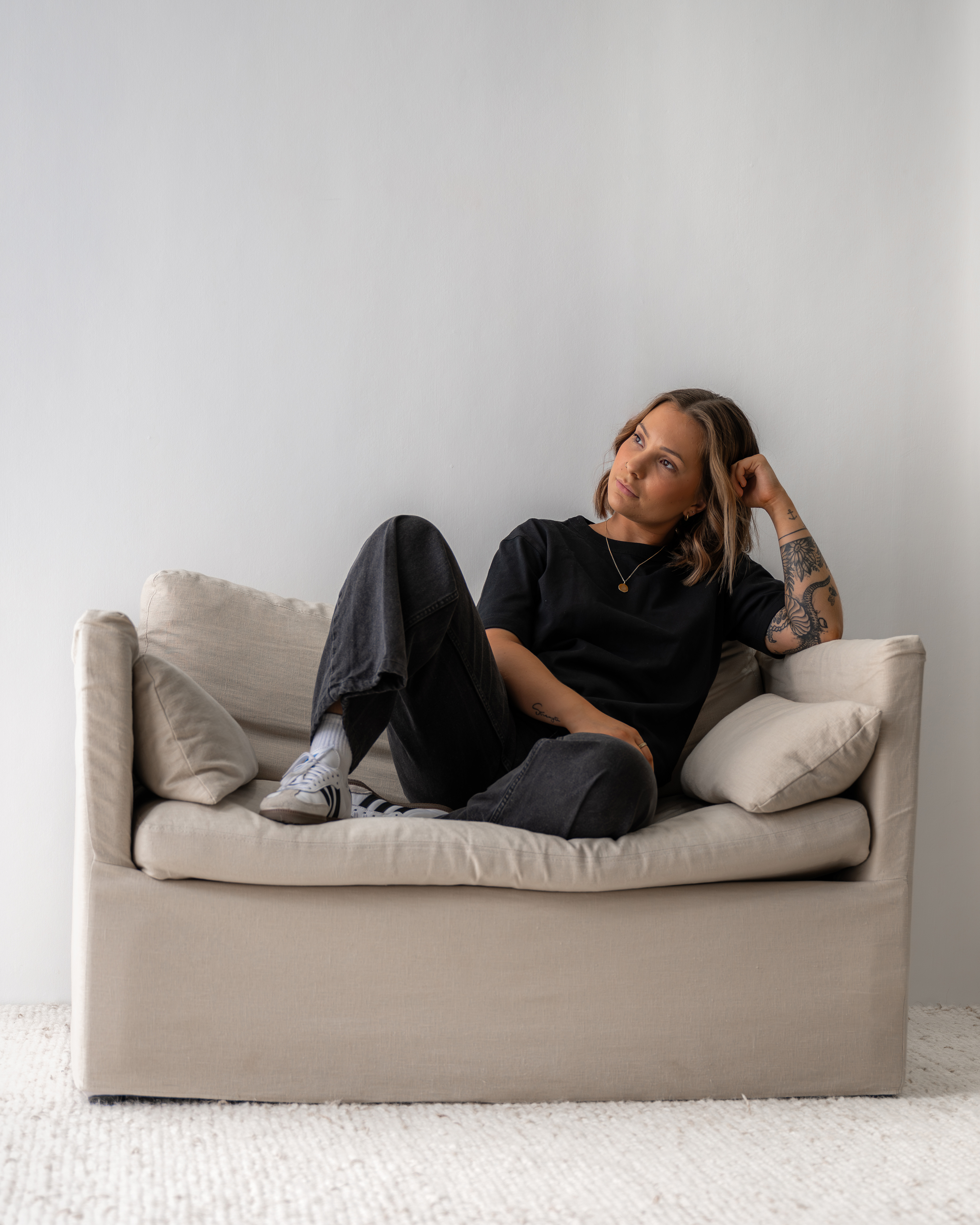 Young woman with tattoos sitting casually on a beige sofa against a plain white wall, looking thoughtfully to the side.