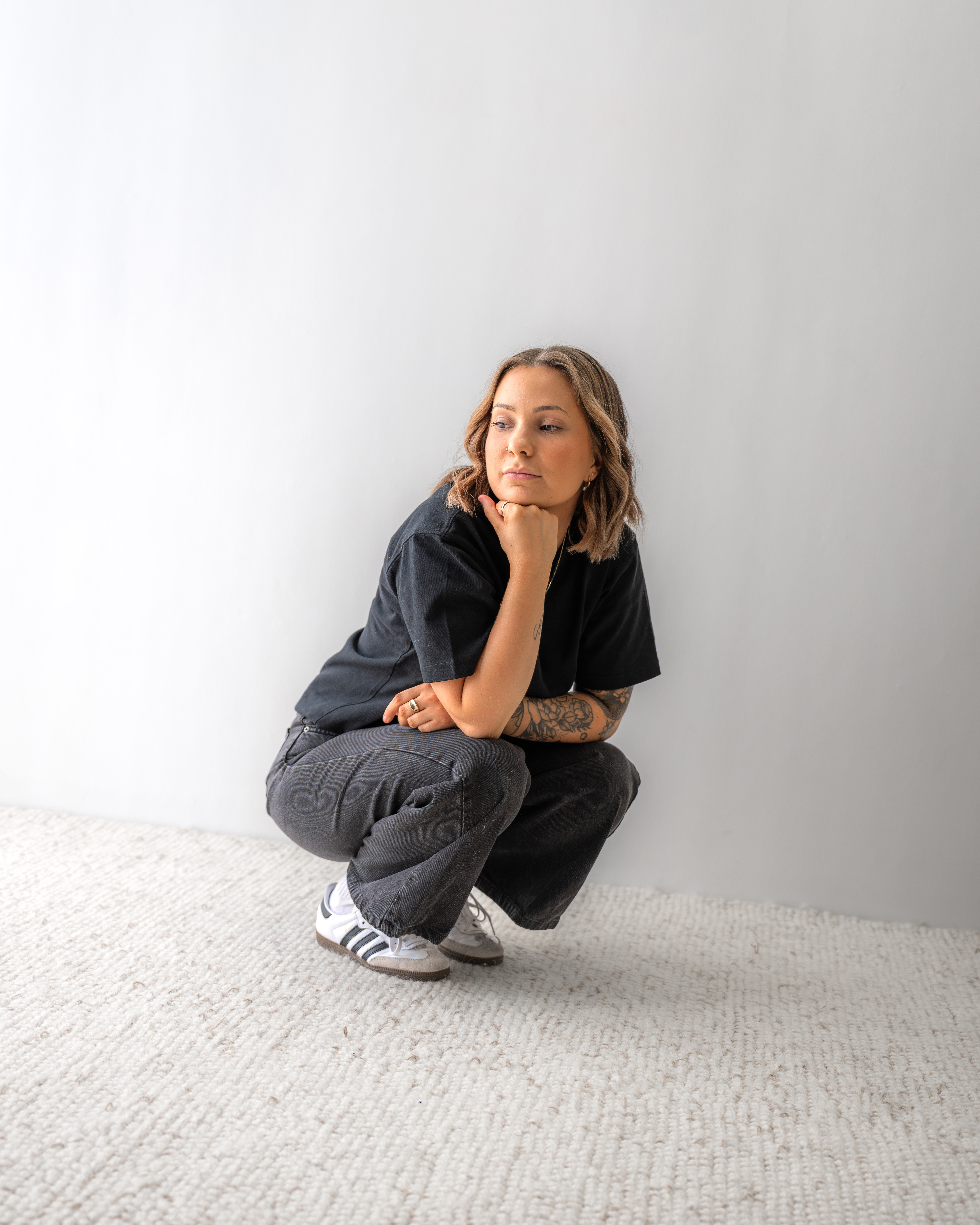 Woman with tattoos squatting on a cream carpet against a plain white wall, resting her chin on her hand and looking to the side.