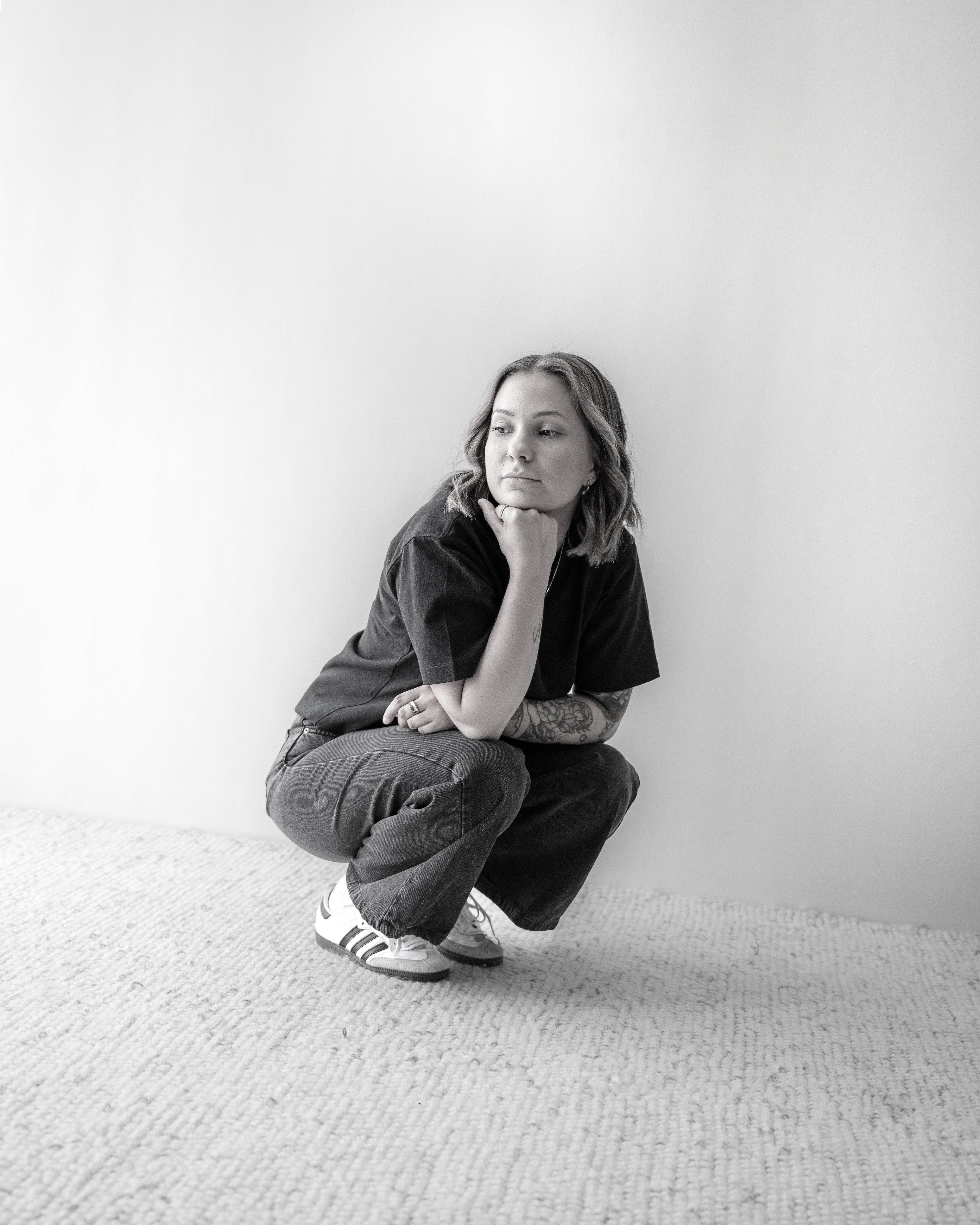 Woman with short hair, tattoos, and casual clothes squatting on a textured carpet against a plain wall, looking to the side thoughtfully.