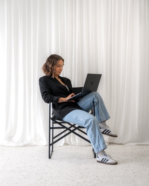 Woman in black blazer and blue jeans sitting on a black folding chair using a laptop, with white curtains in the background.