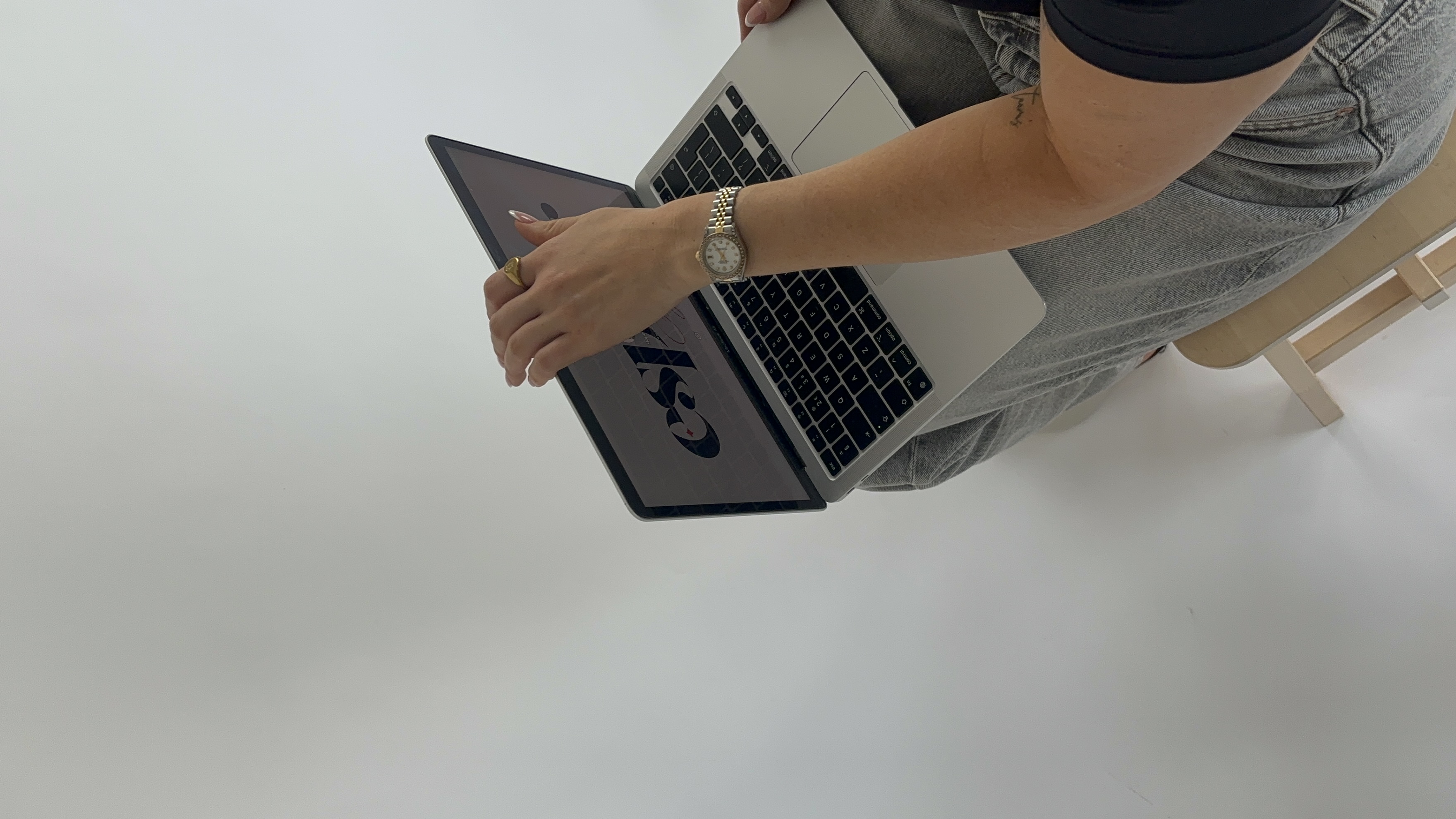 Person wearing a watch and ring holding a laptop displaying a design with the text 'csp' while sitting on a wooden bench.