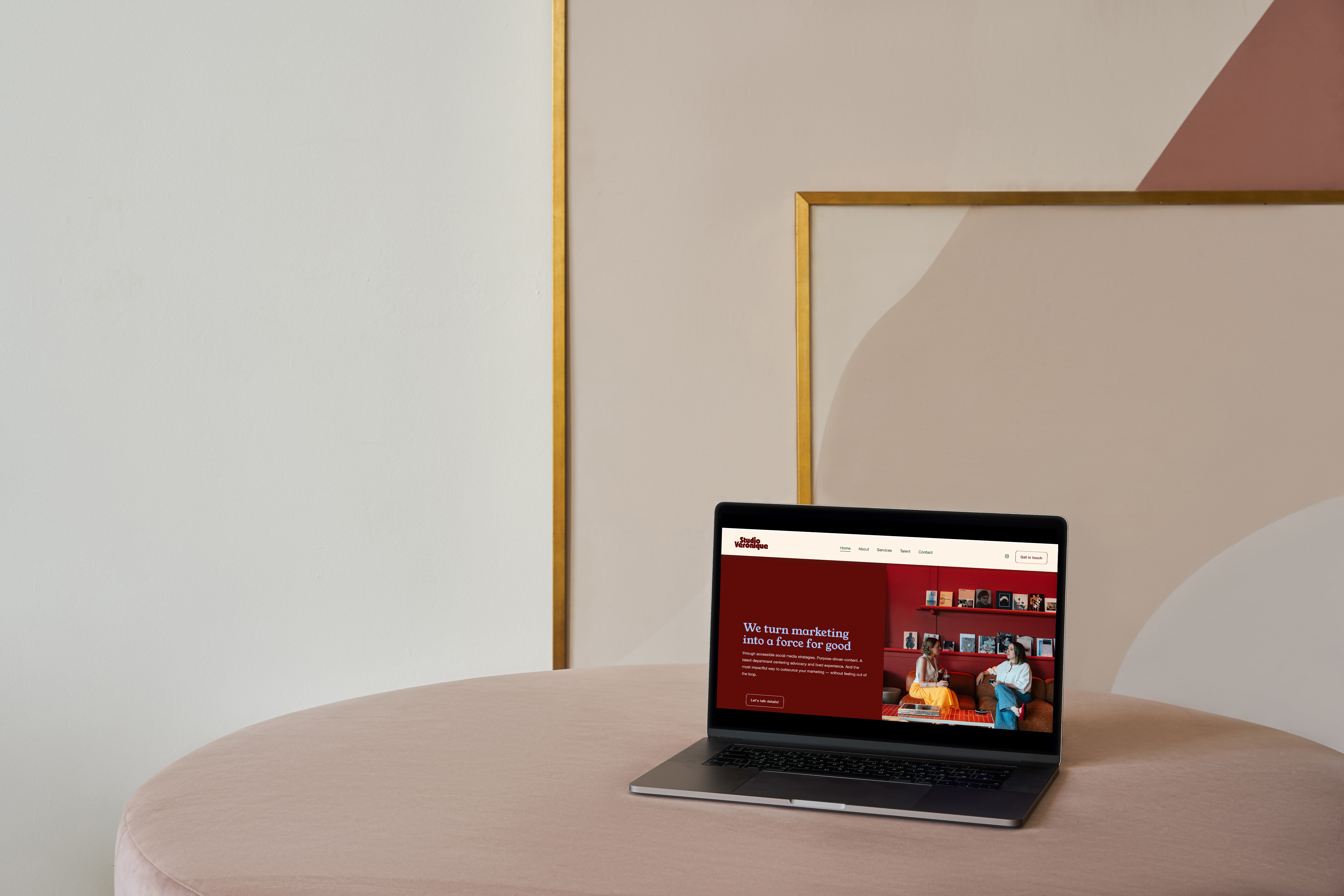 Open laptop on a round pink table displaying a website with the message 'We turn marketing into a force for good' and an image of two women talking on a red couch.