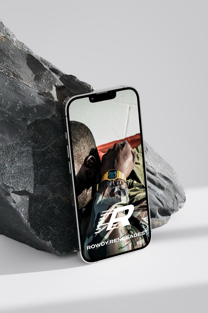 Smartphone leaning against a textured rock displaying a man wearing a camouflage jacket and a gold watch with the text 'Rowdy Renegades' and a stylized R logo.