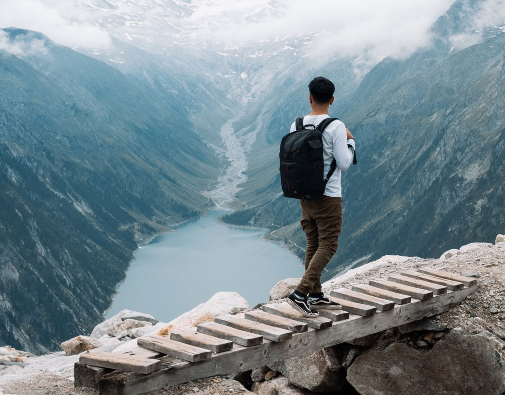 Man walking on a wooden bridge on top of a mountain overlooking a valley with a lake