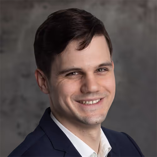 Porträt eines lächelnden jungen Mannes mit dunklen Haaren, der einen dunklen Anzug und ein weißes Hemd trägt, vor einem grauen Hintergrund.