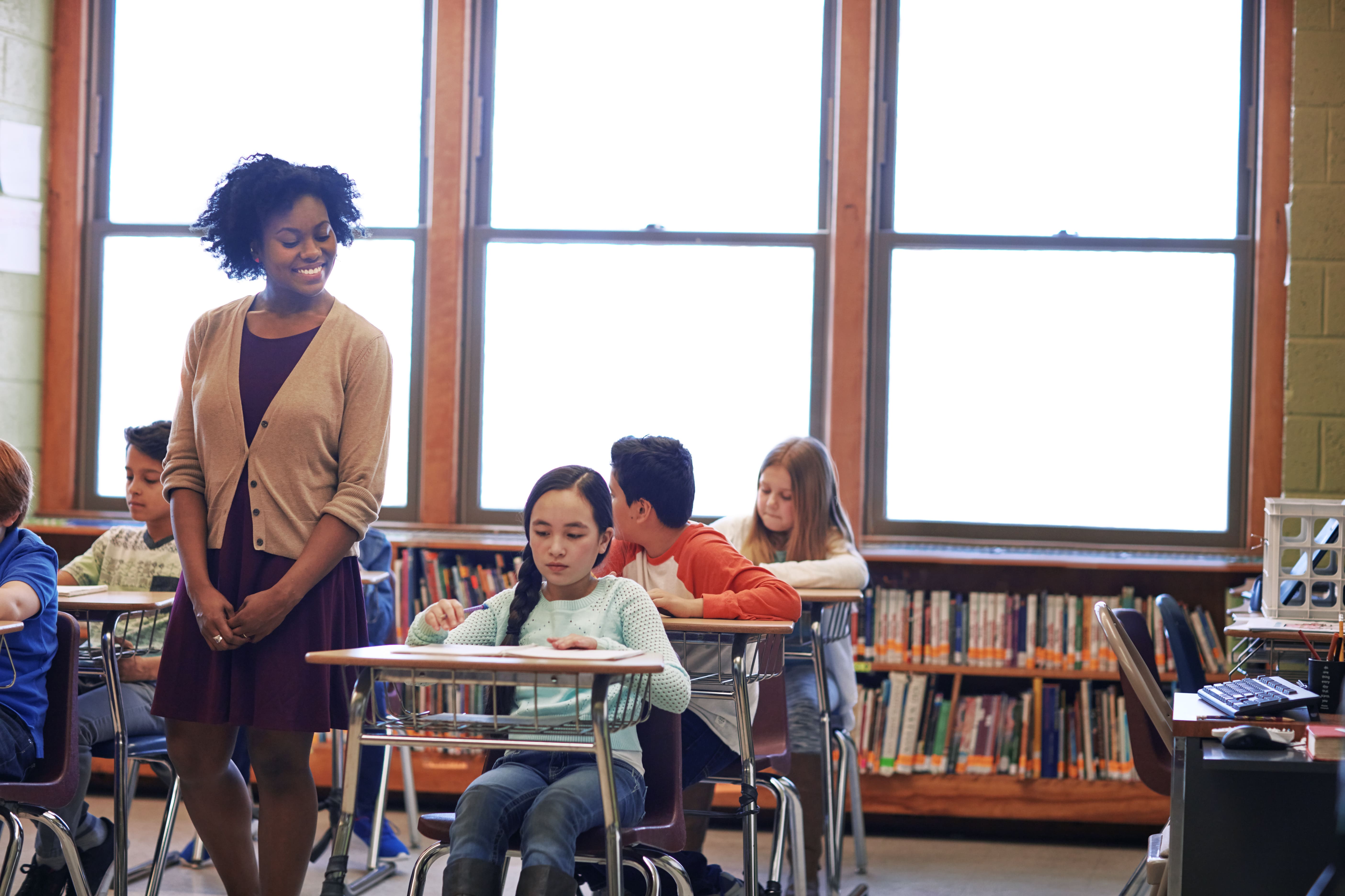 A female teacher standing attentively by students' desks in a bright, diverse public school classroom, emphasizing a supportive learning environment.