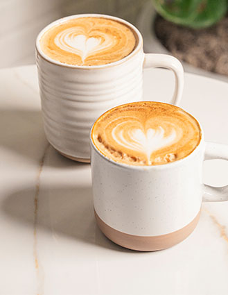 Two white mugs of latte with heart-shaped foam art on a white marble surface.
