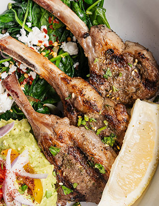 Grilled lamb chops served with sautéed spinach, crumbled cheese, guacamole, and a lemon wedge.