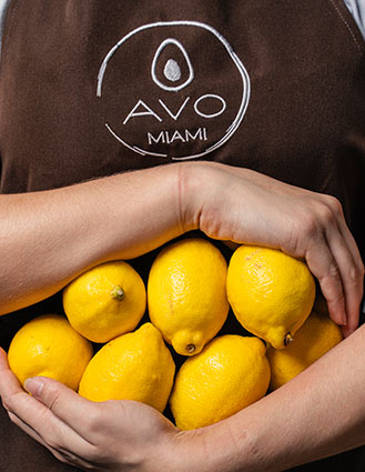 Person wearing an AVO Miami apron holding several bright yellow lemons close to their chest.
