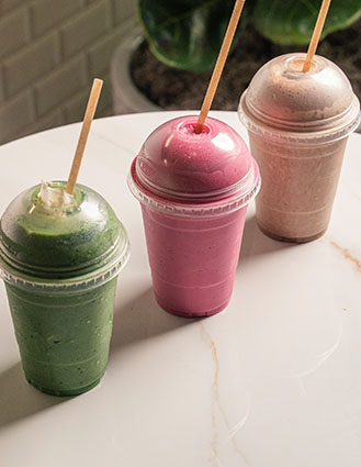 Three plastic cups with dome lids containing green, pink, and beige smoothies with wooden straws on a white round table.