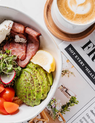 Bowl with sliced avocado, tomato, radish, microgreens, bacon, and a lemon wedge next to a cup of coffee with latte art on a wooden coaster.